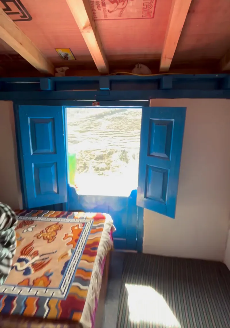 Upstairs guest room with two beds, colourful bedcovers, wooden beams and a blue window