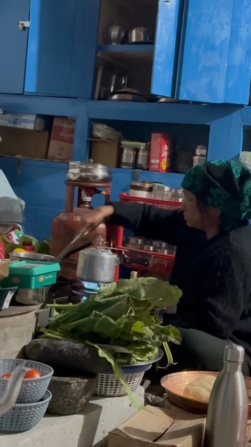 Mati at her gas stove with a pressure cooker and a pile of fresh greens